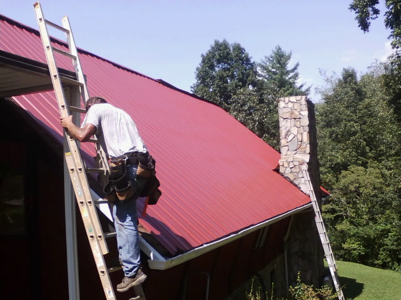 Roofer on a ladder beside a red metal roof and stone chimney — Soffit & Fascia Repair by Roofing Brothers in Lookeba